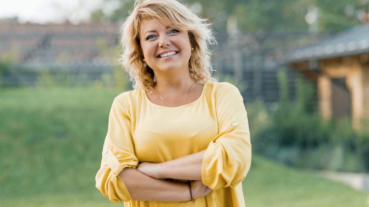 Smiling female blonde in a yellow dress with arms crossed near the house